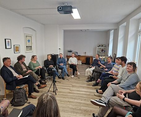 Ausschnitt aus der Gesprächsunde. Eine Gruppe von Menschen sitzt in einem Kreis und hören einer erzählenden Person (3.v.r.) zu. 
